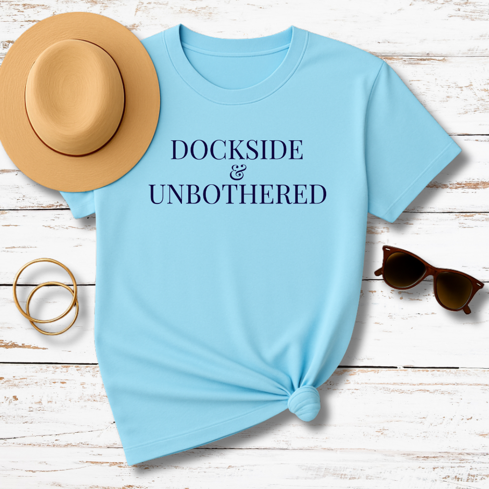 Light blue t-shirt with 'DOCKSIDE & UNBOtherED' text, accompanied by a hat, sunglasses, and rings on a wooden surface.