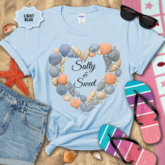 Light blue t-shirt with seashell design and 'Salty & Sweet' text, surrounded by beach accessories on sand.