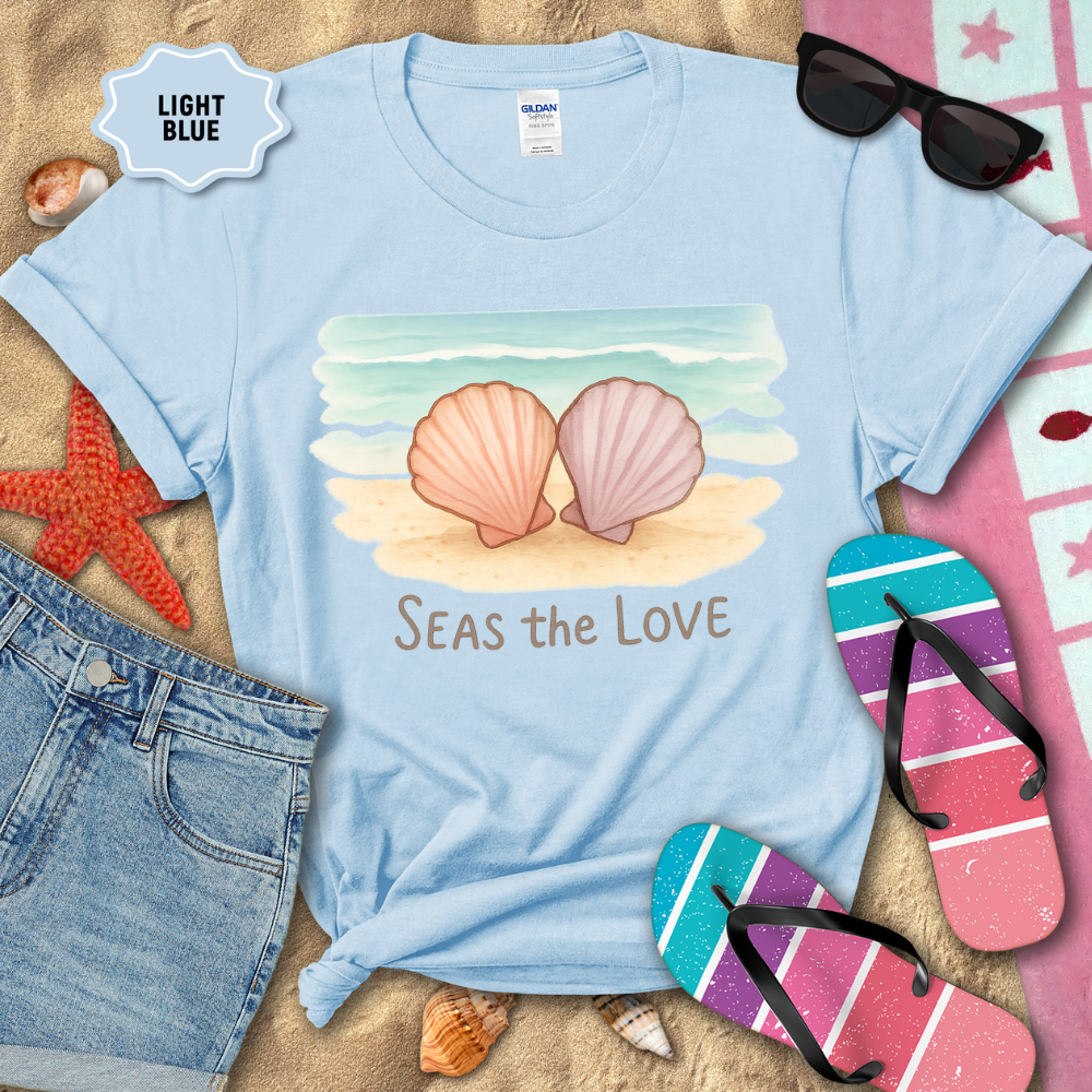 Light blue t-shirt with shell design and 'Seas the Love' text, surrounded by beach accessories on sand.