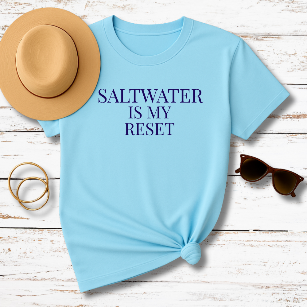 Light blue t-shirt with 'Saltwater is my reset' text, placed on a wooden surface with a hat, sunglasses, and rings.