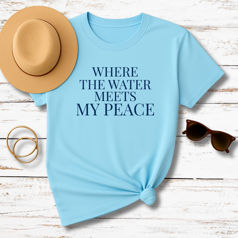 Light blue t-shirt with 'WHERE THE WATER MEETS MY PEACE' text, placed on a wooden surface with a hat, sunglasses, and rings.
