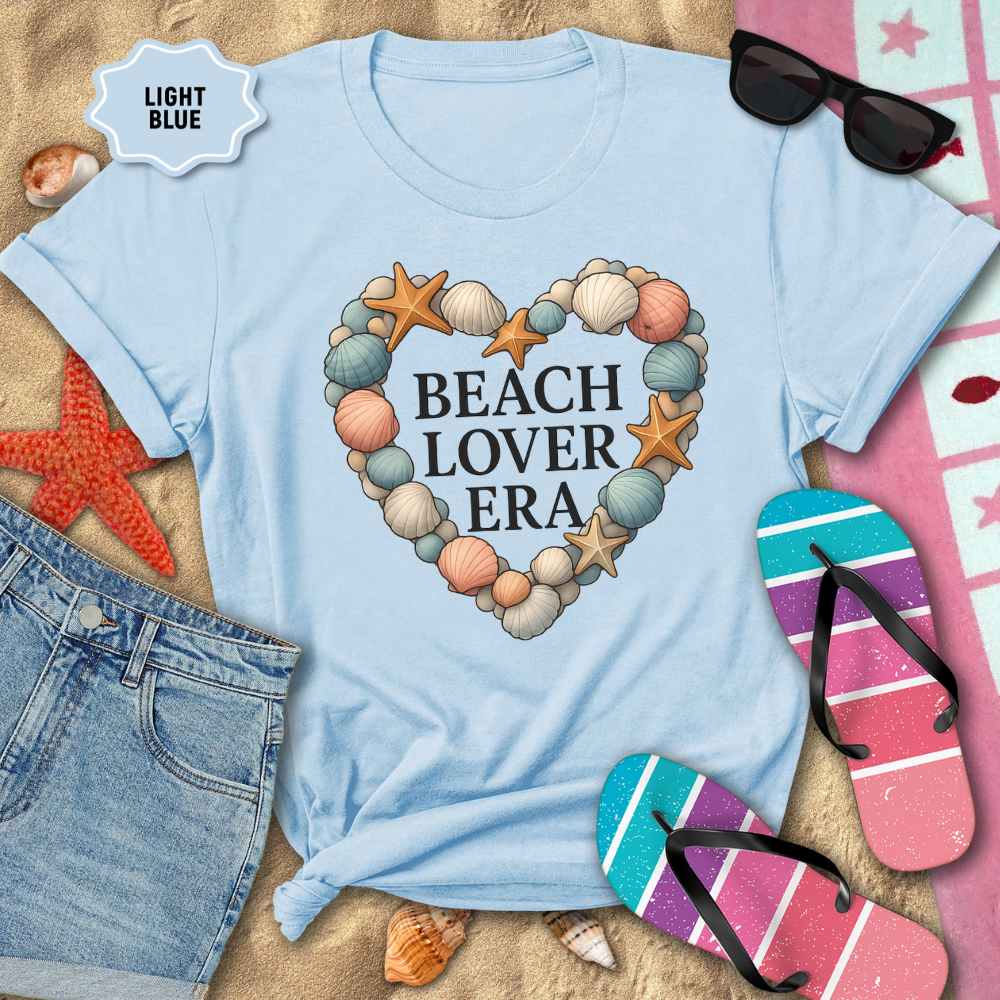 Light blue t-shirt with 'Beach Lover Era' design on sand with beach accessories.