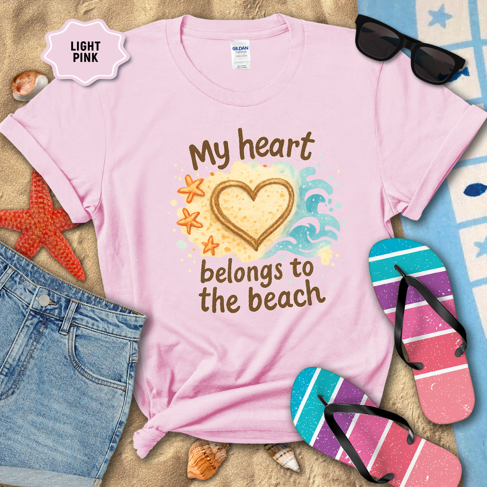 Light pink t-shirt with 'My heart belongs to the beach' design on a sandy background with flip-flops and sunglasses.