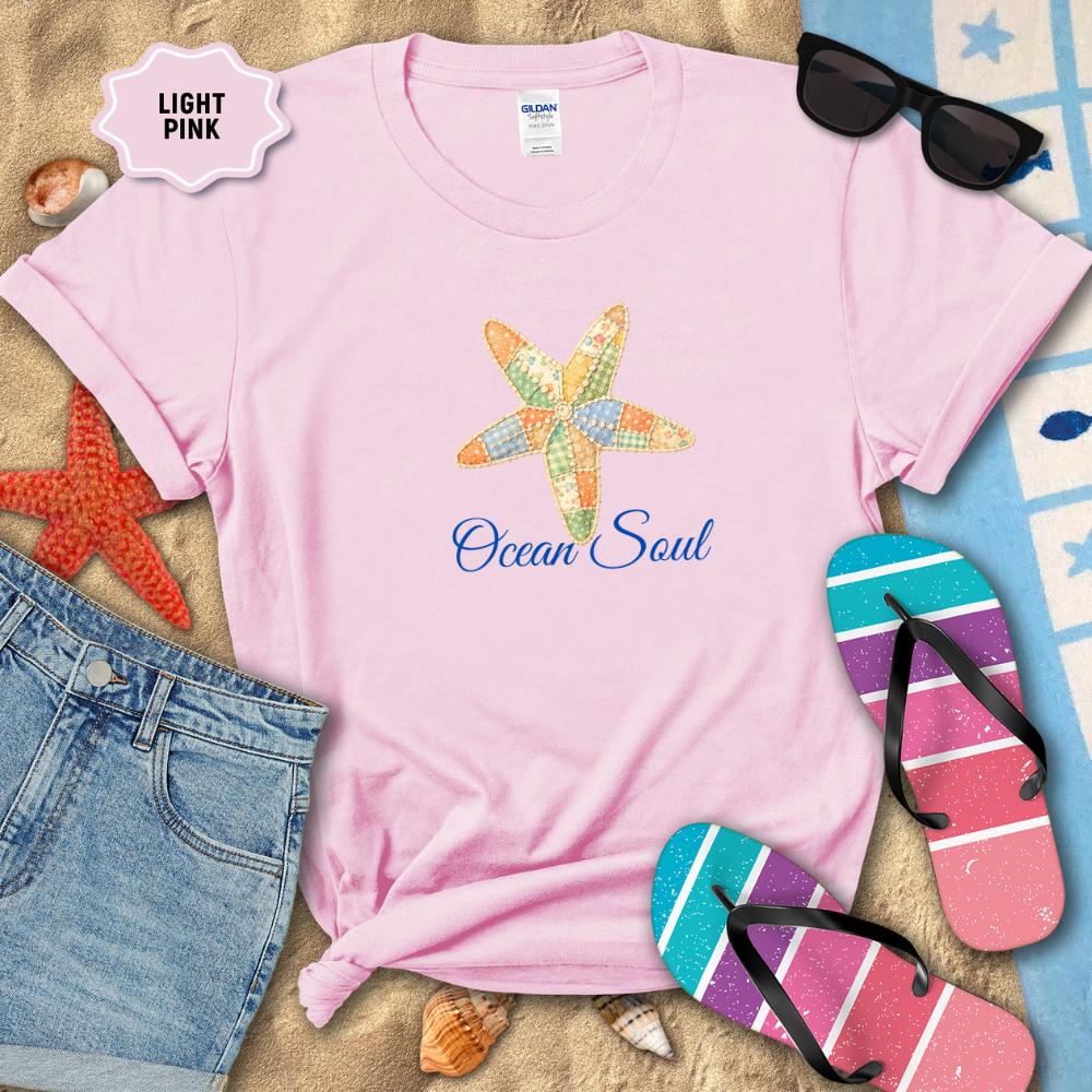 Light pink t-shirt with starfish design and 'Ocean Soul' text, surrounded by beach accessories on sand.