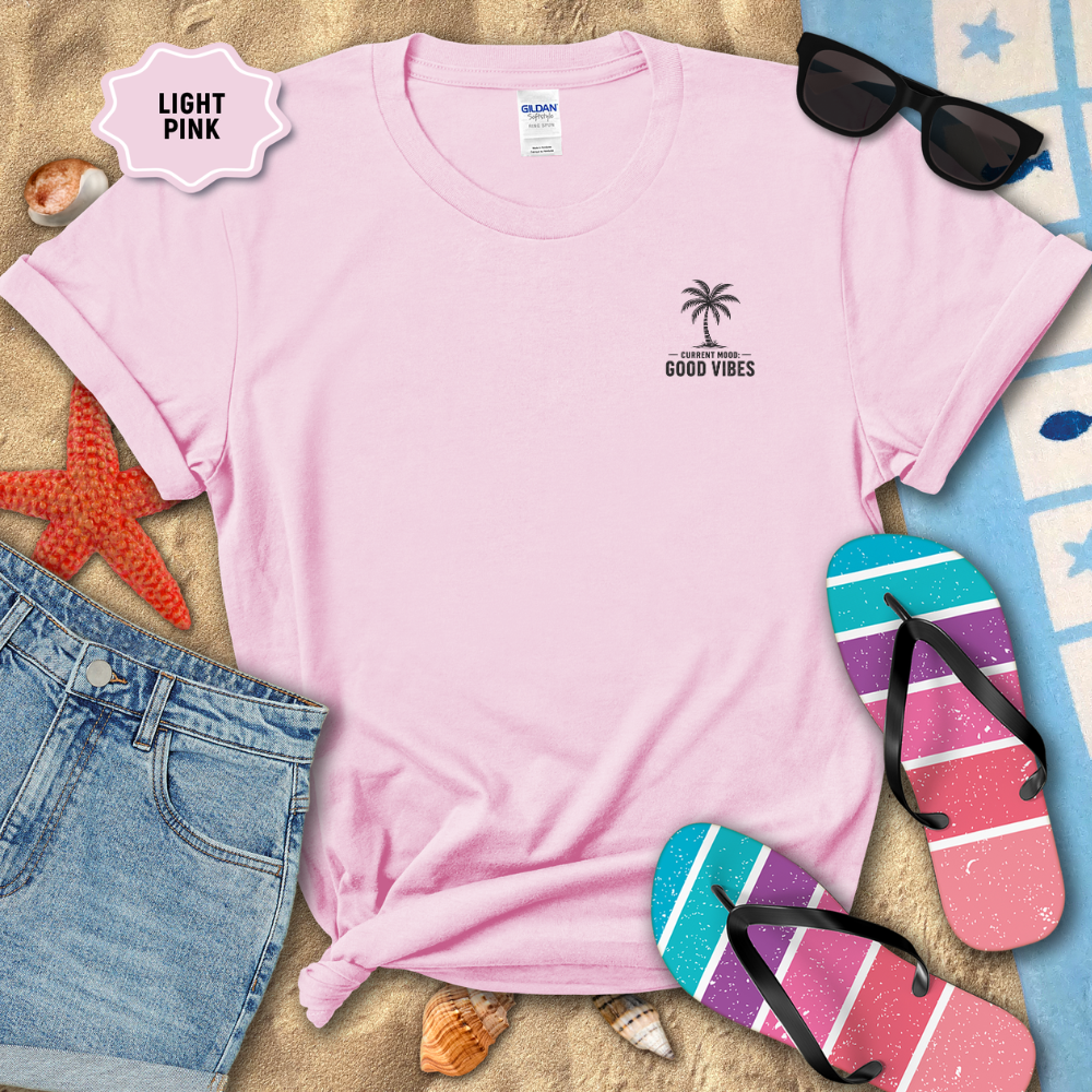 Light pink t-shirt with palm tree design and 'Good Vibes' text, surrounded by beach accessories on sand.