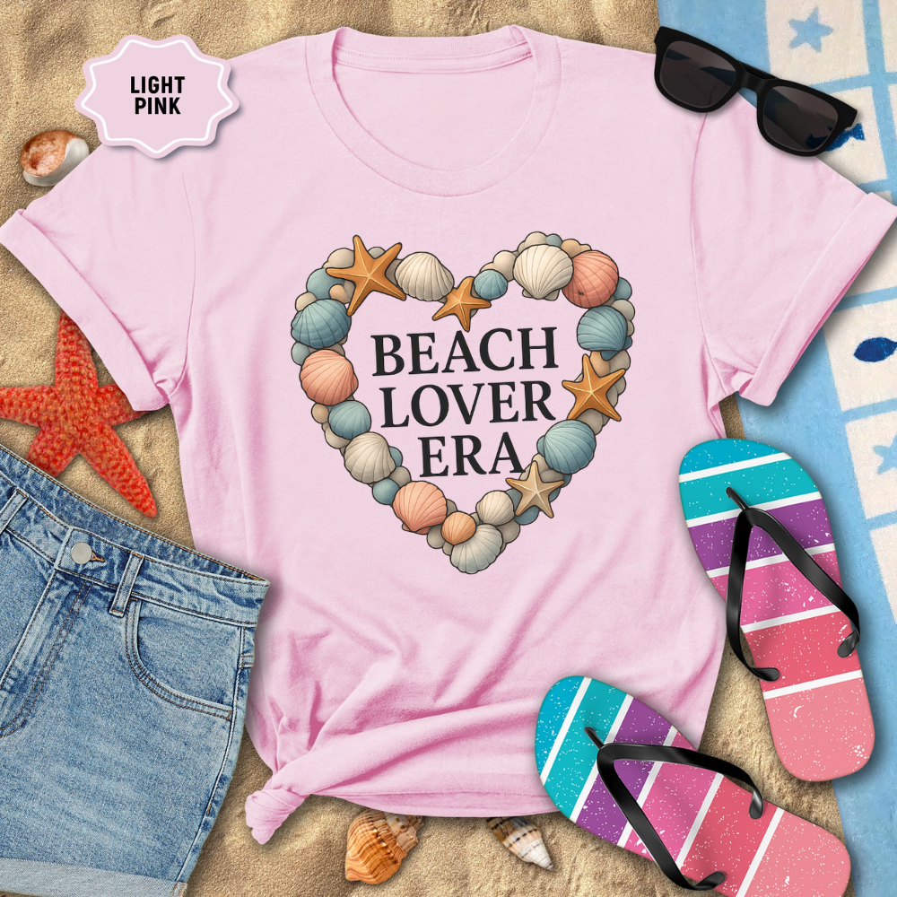 Light pink t-shirt with 'Beach Lover Era' design on a sandy background with beach accessories.
