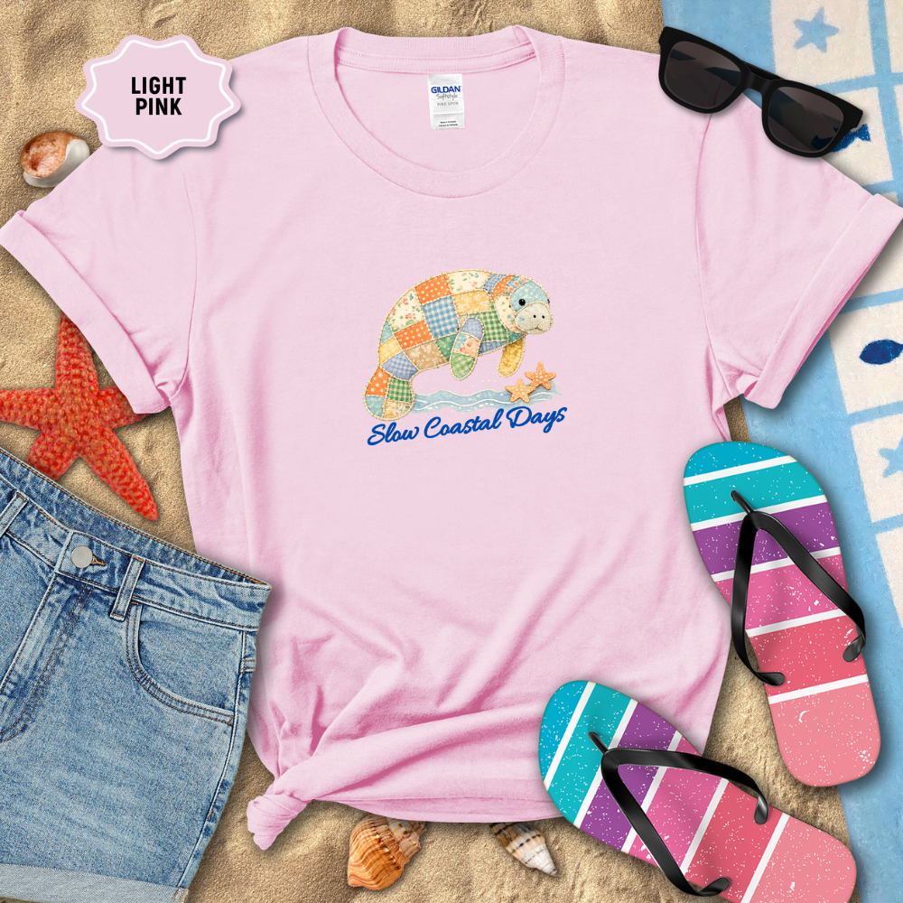 Light pink t-shirt with turtle design and text, surrounded by beach items on sand.