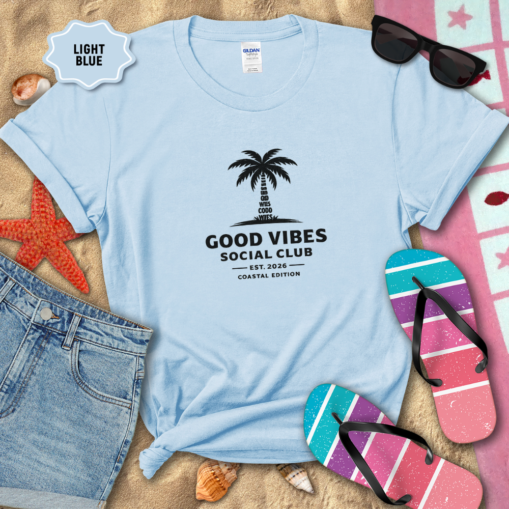 Light blue t-shirt with 'Good Vibes Social Club' design on sand with beach accessories.