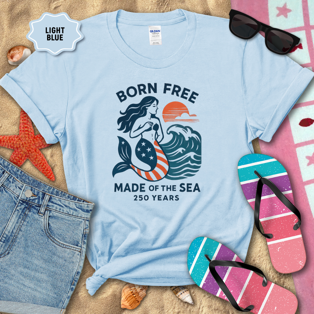 Light blue t-shirt with mermaid graphic and text on a sandy background with beach accessories.