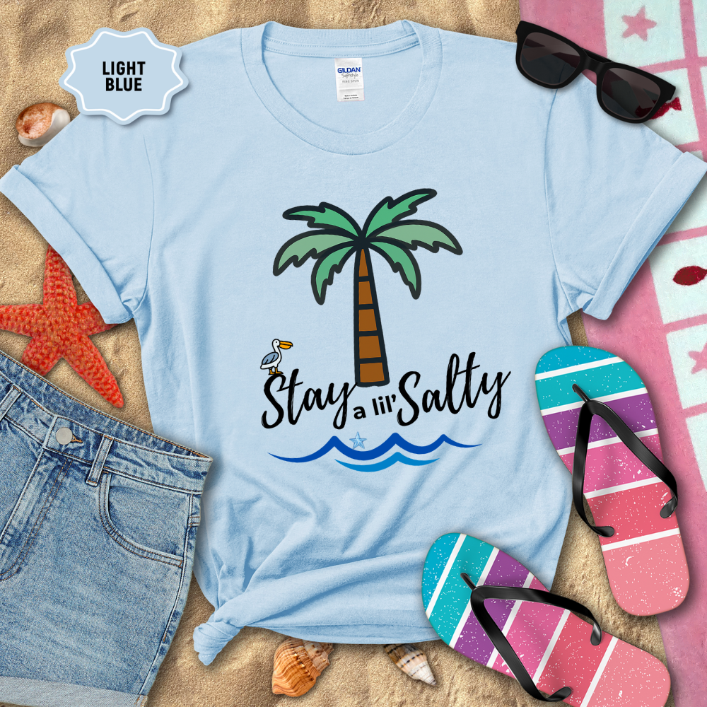Light blue t-shirt with palm tree and 'Stay a lil' Salty' text, surrounded by beach accessories on sand.