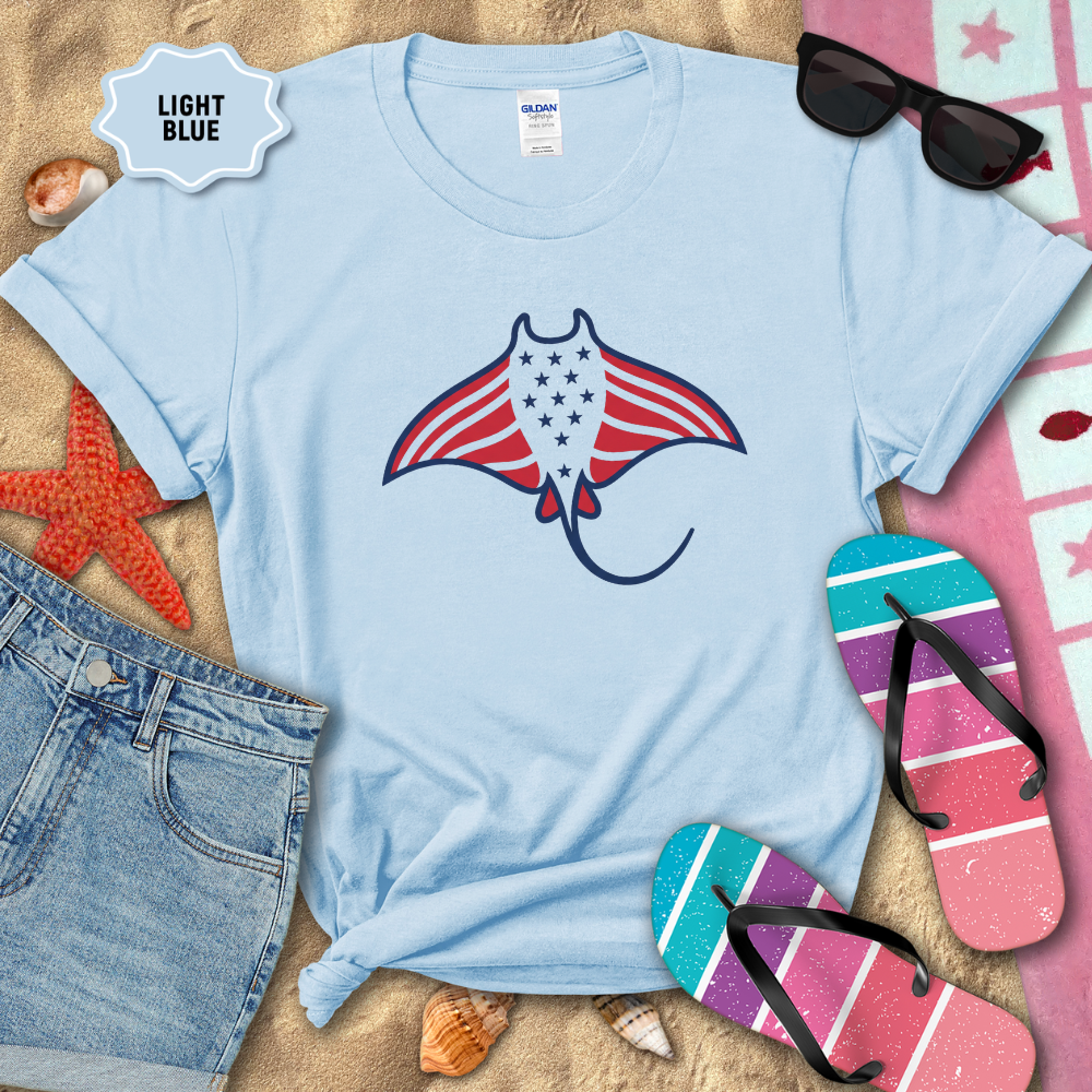 Light blue t-shirt with a graphic of a ray fish with an American flag design, surrounded by beach items on sand.