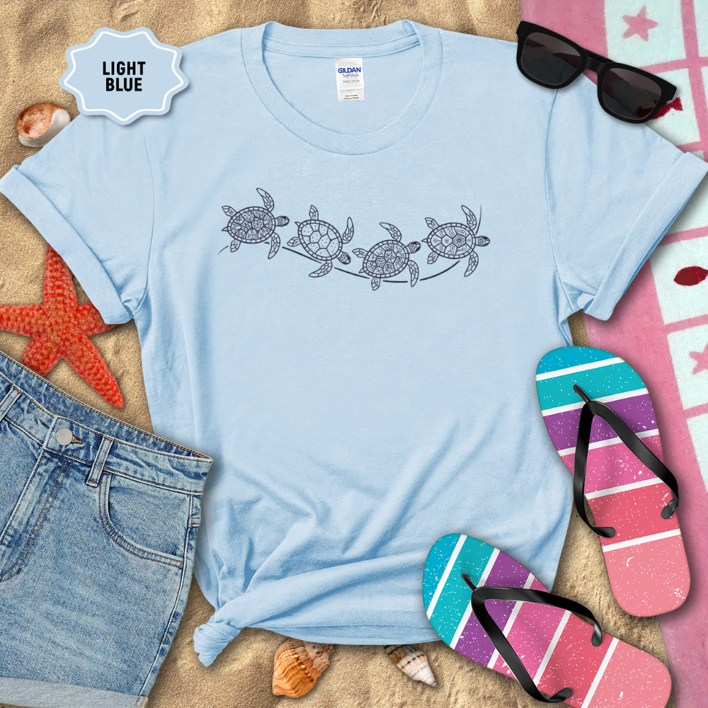 Light blue t-shirt with turtle design on a sandy background with beach accessories.