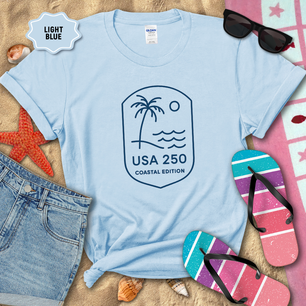 Light blue t-shirt with a palm tree and wave design, surrounded by beach accessories on sand.