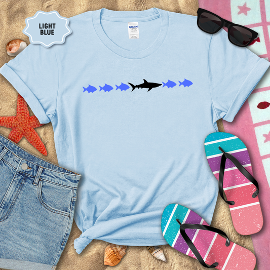 Light blue t-shirt with shark and fish design on a sandy background with flip-flops and sunglasses.