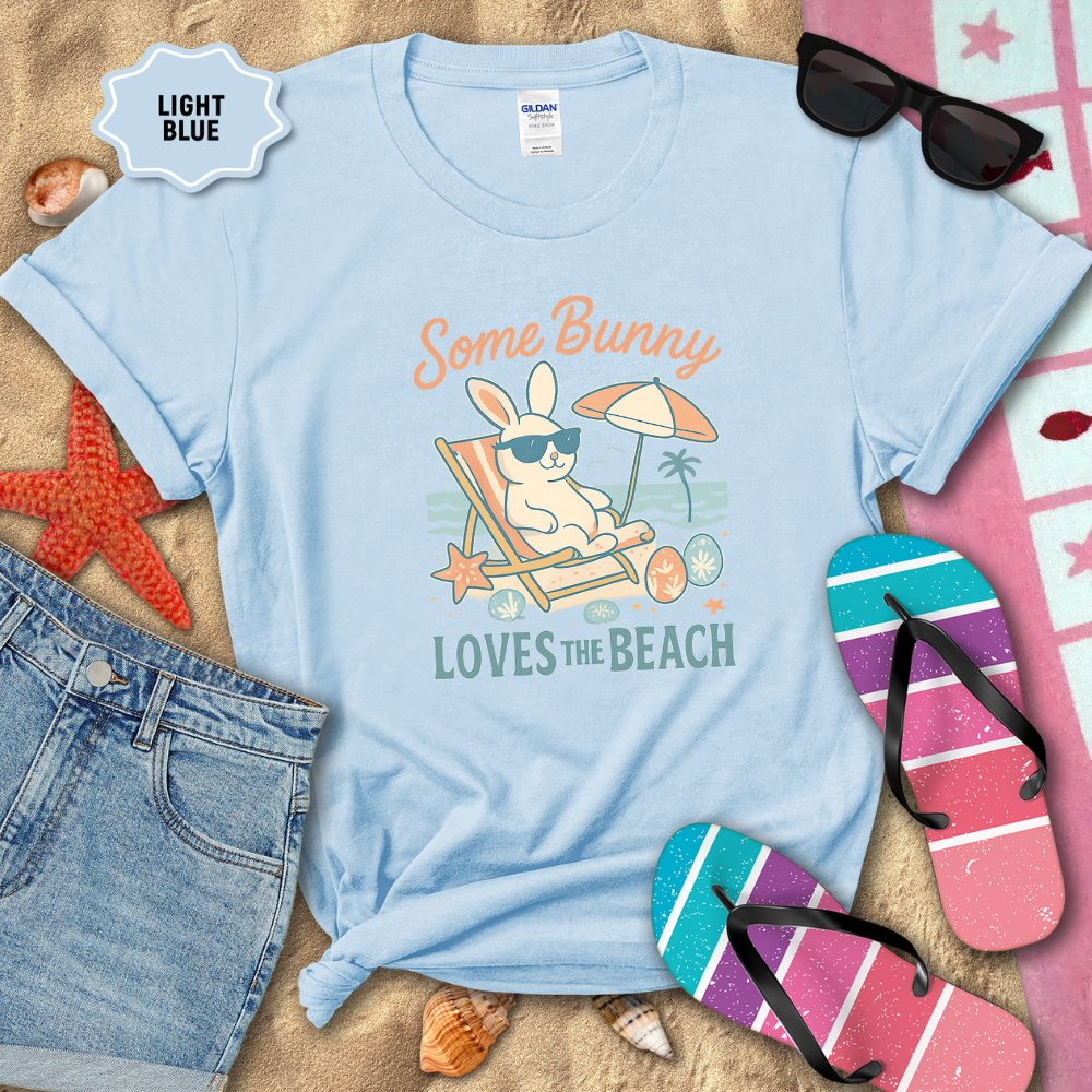 Light blue t-shirt with beach-themed design featuring a bunny, sunglasses, and umbrella, surrounded by beach accessories on sand.
