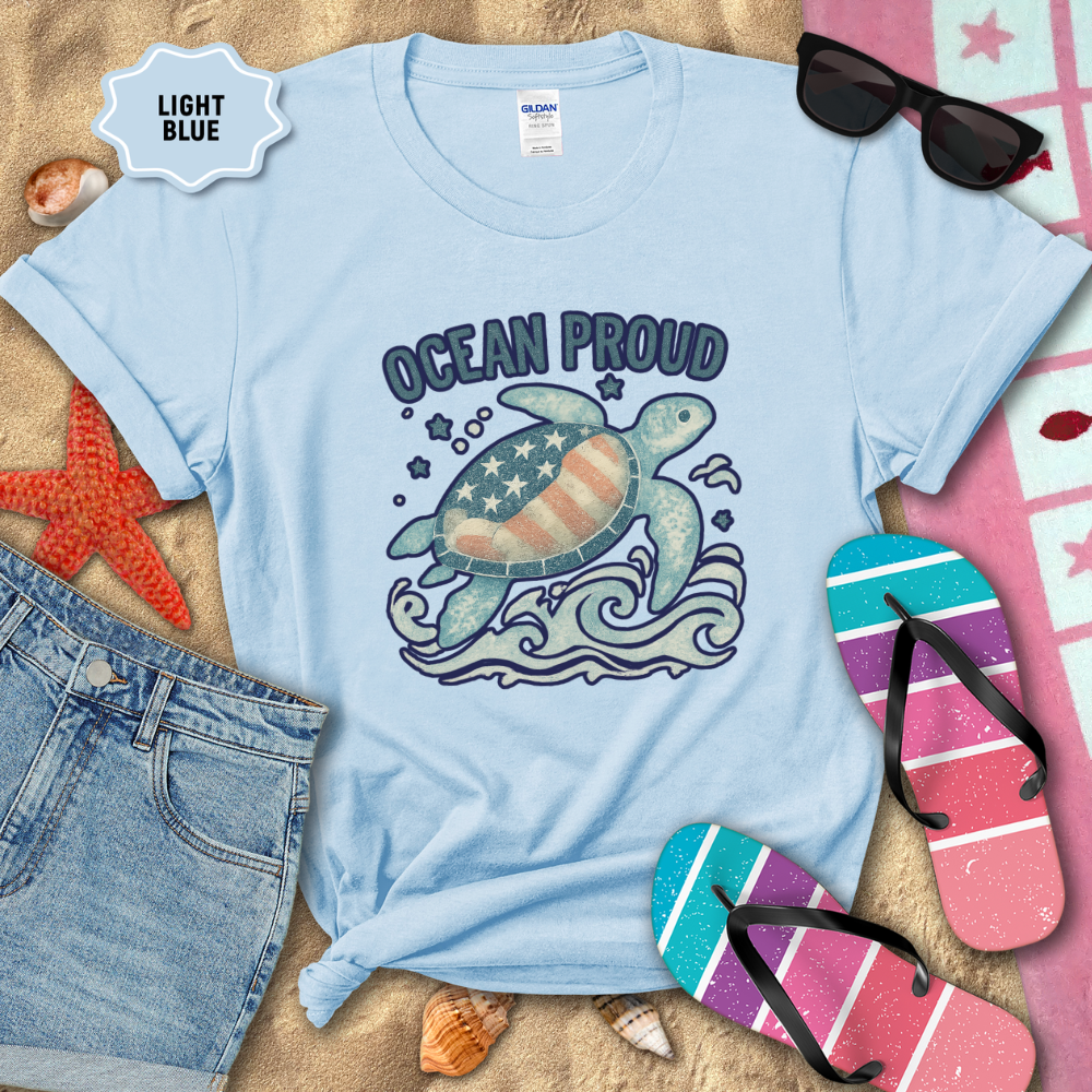 Light blue t-shirt with turtle graphic and 'Ocean Proud' text on a sandy background with beach accessories.