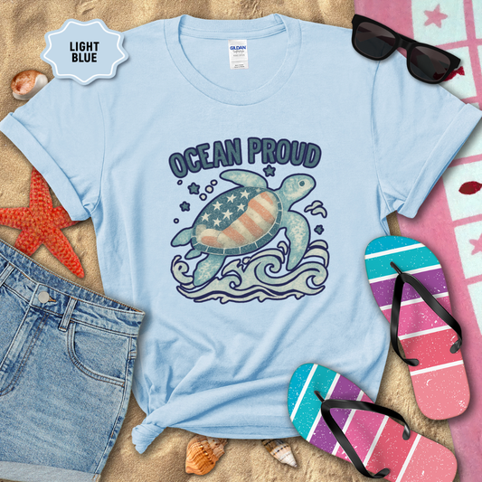 Light blue t-shirt with turtle graphic and 'Ocean Proud' text on a sandy background with beach accessories.
