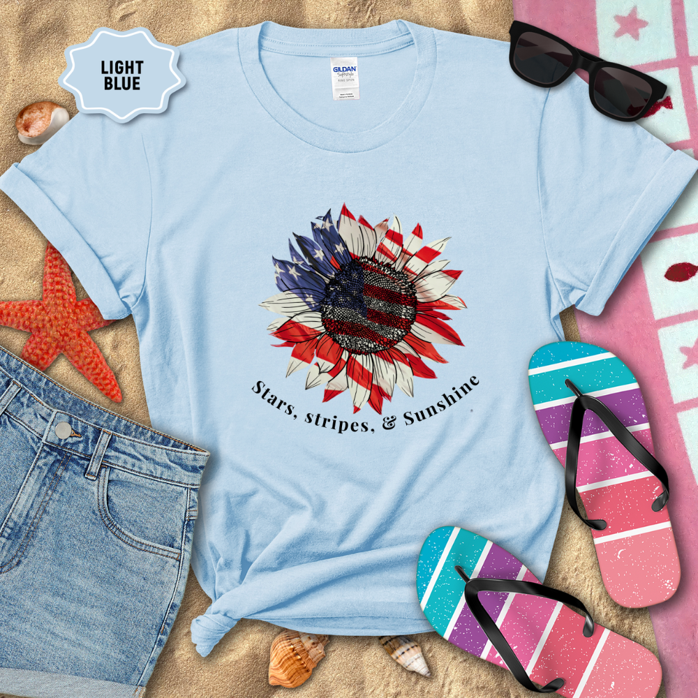 Light blue t-shirt with sunflower design and text on a sandy background with beach accessories.