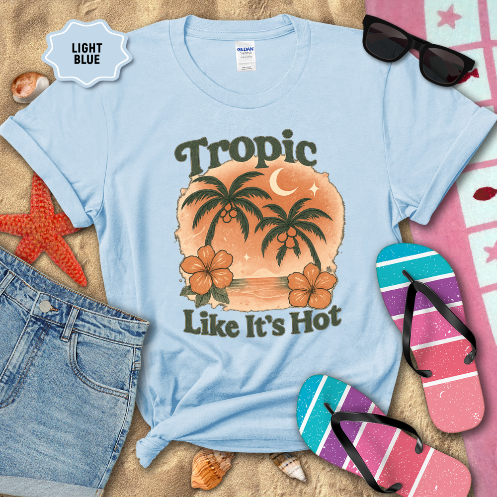 Light blue t-shirt with tropical design and text, surrounded by beach accessories on sand.