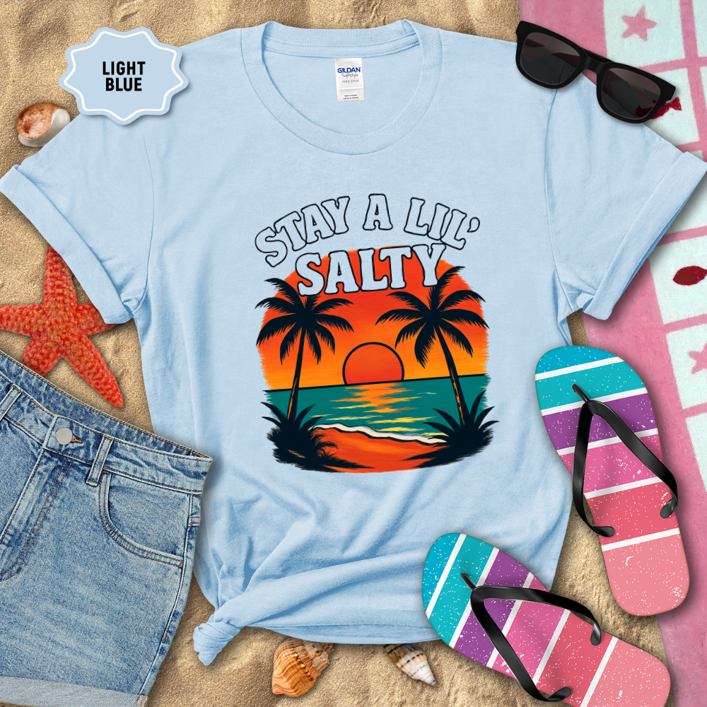 Light blue t-shirt with 'Stay a Lil' Salty' design on sand with beach accessories.
