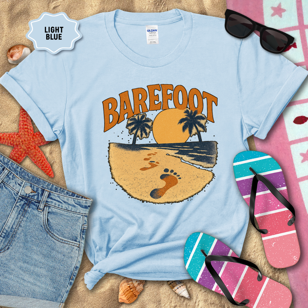 Light blue t-shirt with 'Barefoot' design on sand with beach accessories.