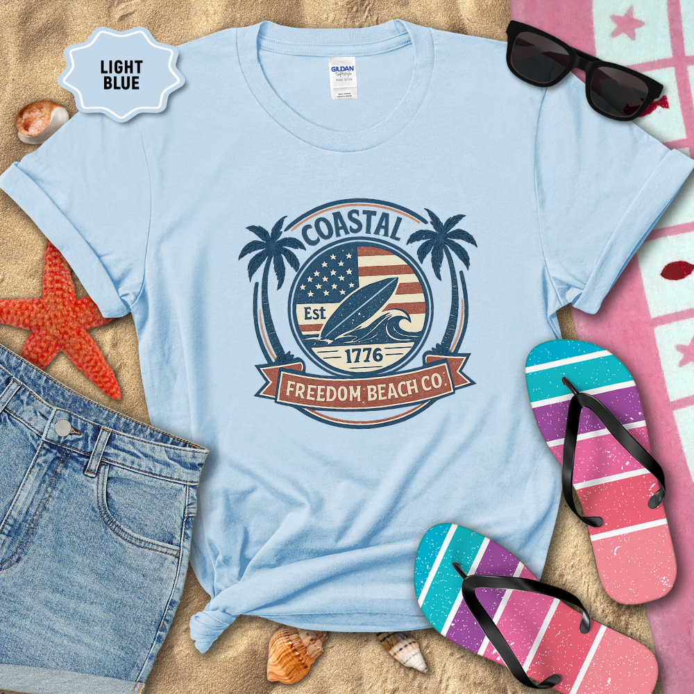 Light blue t-shirt with 'Coastal Freedom Beach Co' design on sand with beach accessories.