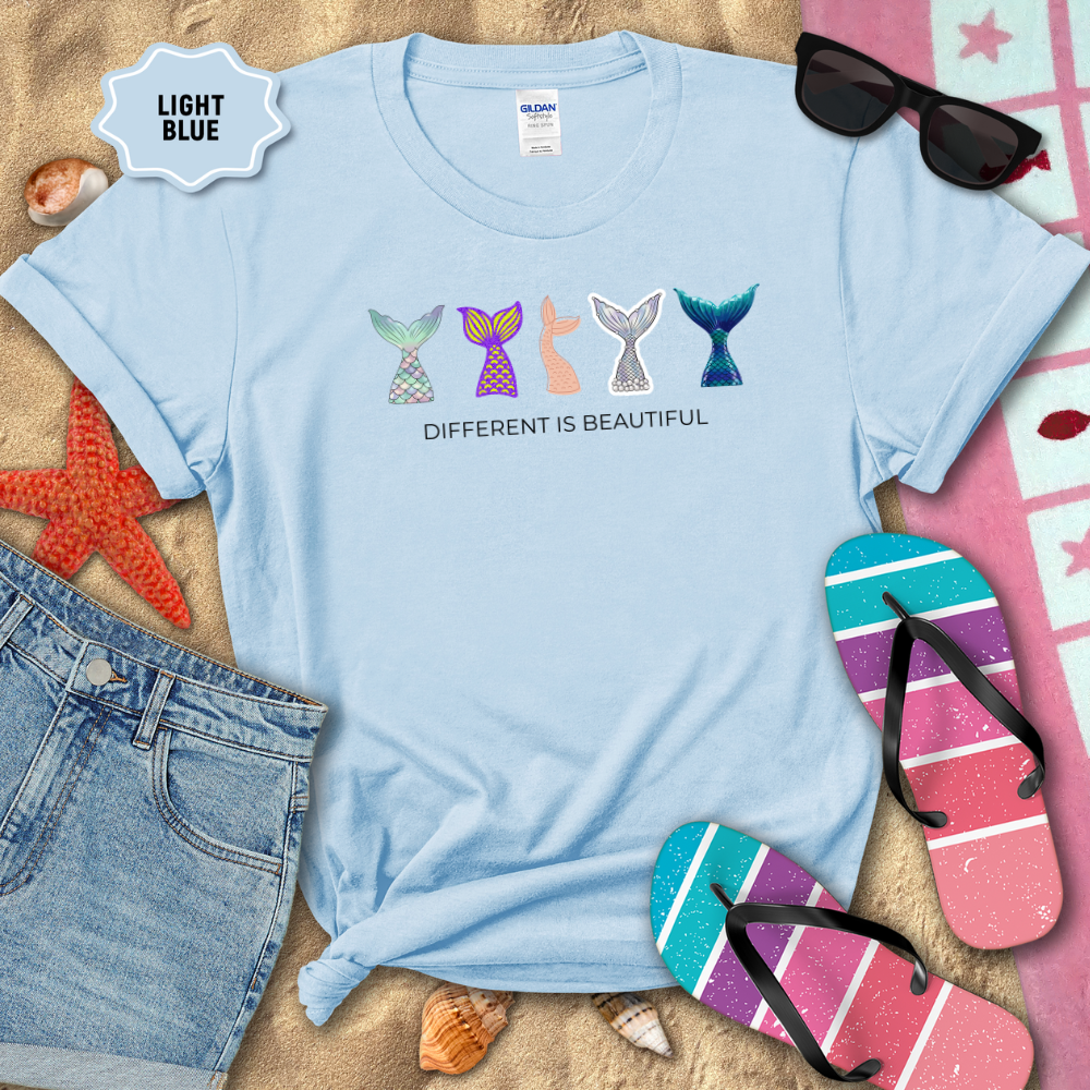 Light blue t-shirt with mermaid tail designs and 'Different is Beautiful' text, placed on sand with beach accessories.