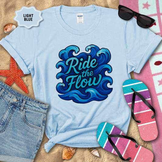 Light blue t-shirt with 'Ride the Flow' design on a sandy background with beach accessories.