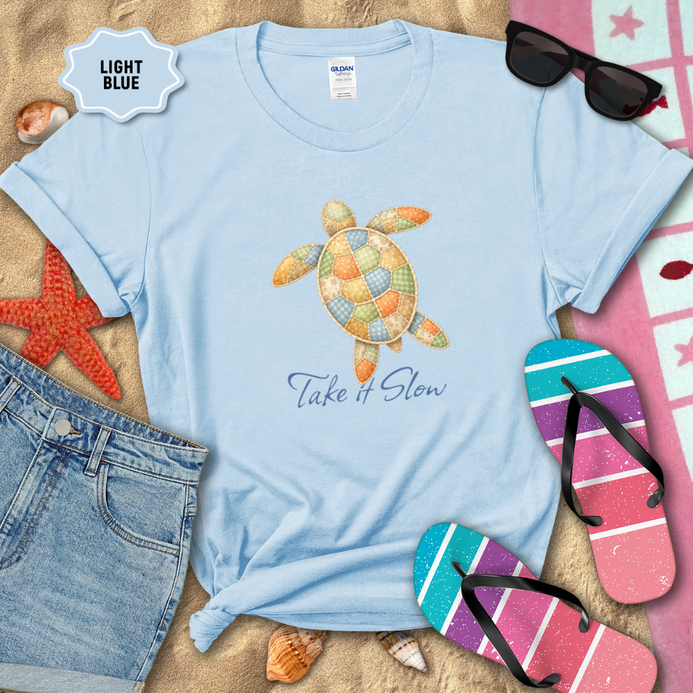 Light blue t-shirt with turtle design and 'Take It Slow' text, surrounded by beach items on sand.