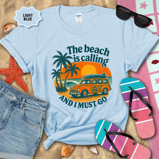 Light blue t-shirt with beach-themed design on sand with flip-flops and sunglasses.