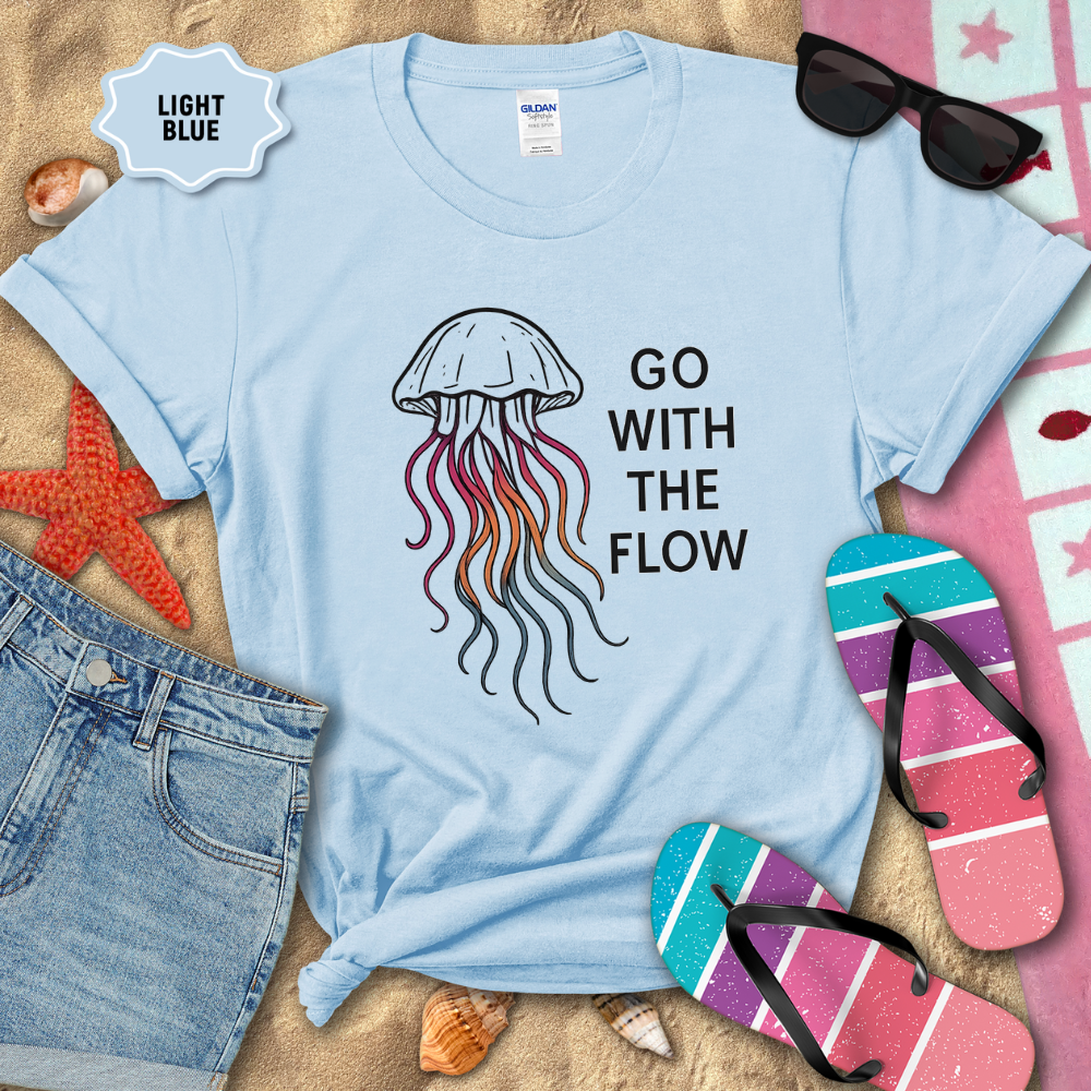 Light blue t-shirt with jellyfish design and 'Go With The Flow' text on a sandy background with beach accessories.