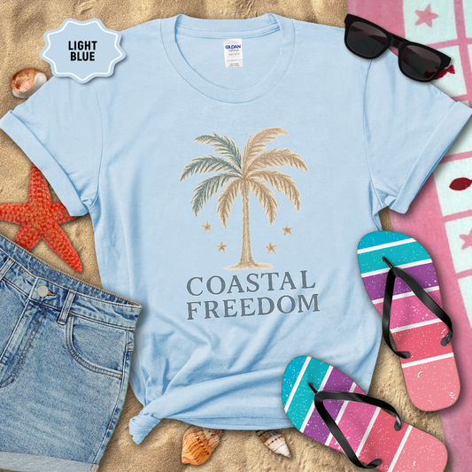 Light blue t-shirt with palm tree design and 'Coastal Freedom' text on a sandy background with beach accessories.