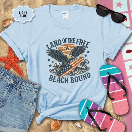 Light blue t-shirt with eagle graphic and 'Land of the Free & Beach Bound' text on a sandy background with beach accessories.