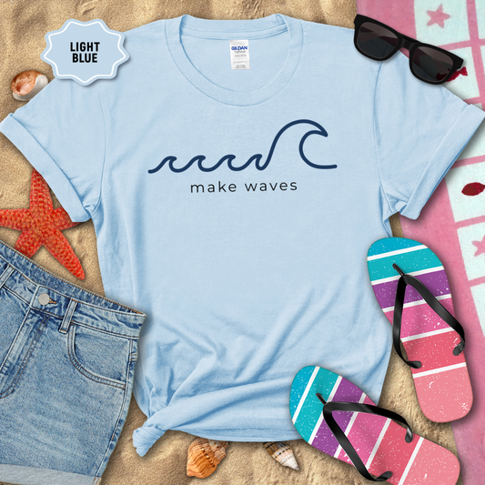 Light blue t-shirt with 'make waves' design on sand with beach accessories