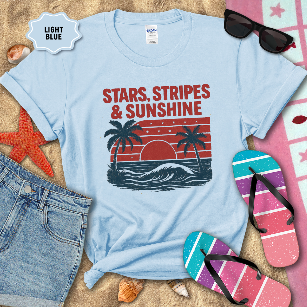 Light blue t-shirt with 'Stars, Stripes & Sunshine' design on a sandy background with beach accessories.