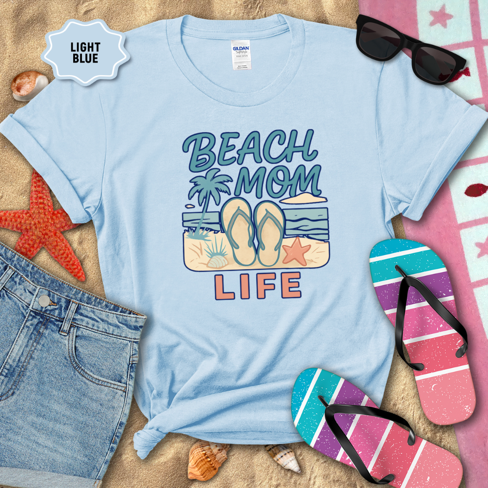 Light blue t-shirt with 'Beach Mom Life' design on sand with beach accessories.