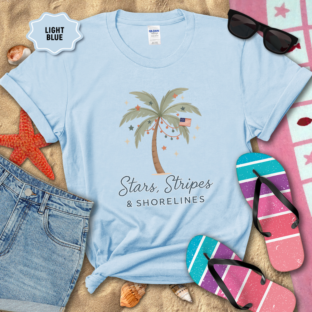 Light blue t-shirt with palm tree and text design on sand with beach accessories.