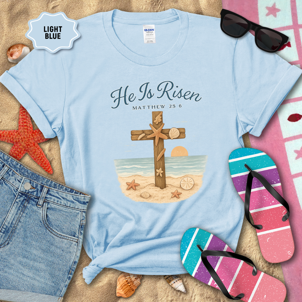 Light blue t-shirt with 'He Is Risen' design on a sandy background with beach accessories.