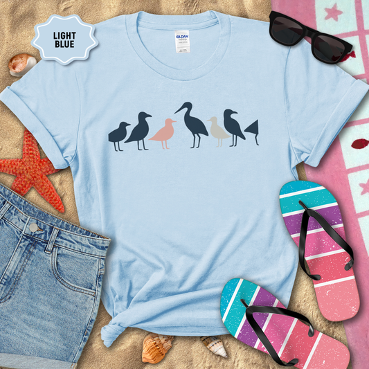 Light blue t-shirt with bird design on a sandy background with beach accessories.