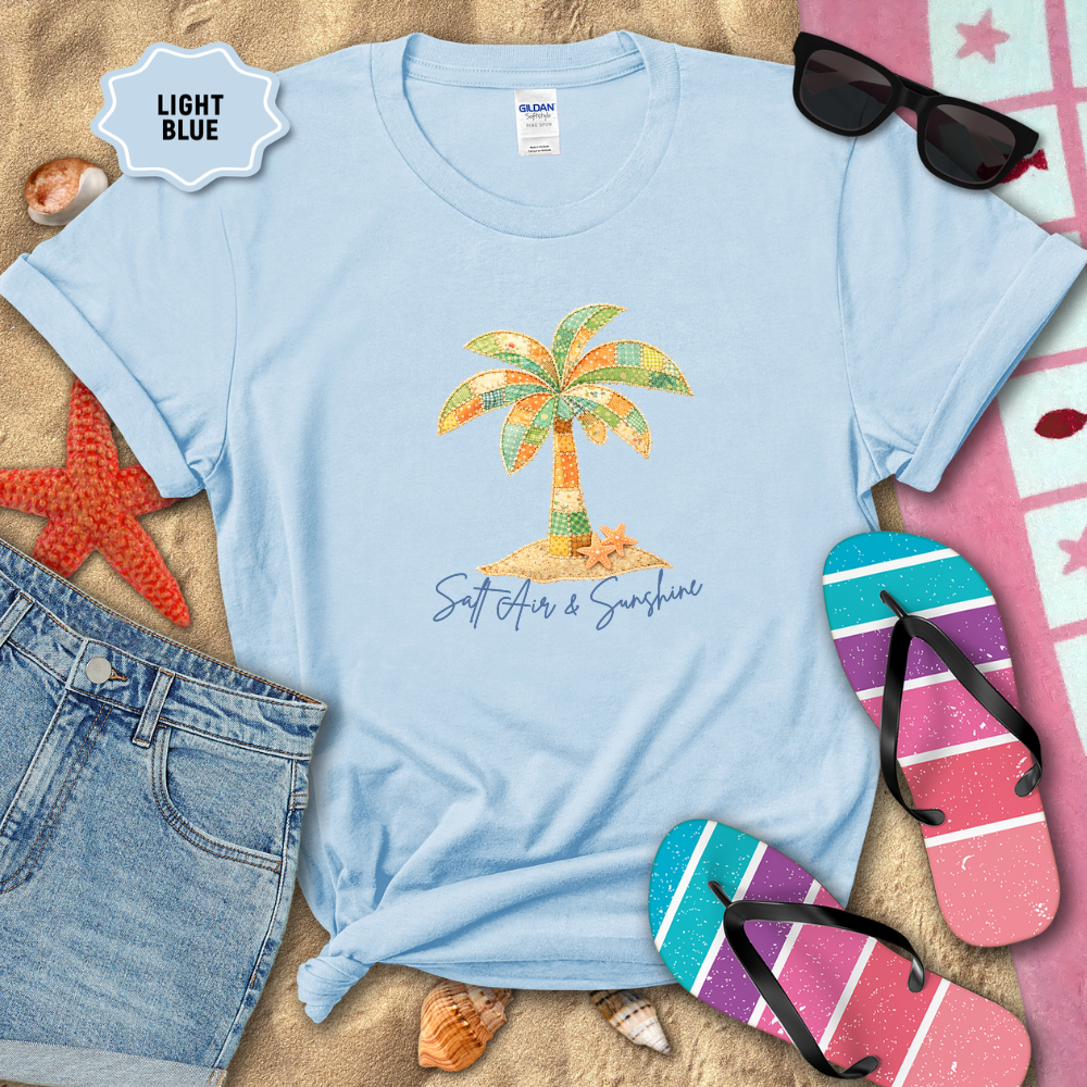 Light blue t-shirt with palm tree design and 'Salt Air & Sunshine' text, surrounded by beach accessories on sand.