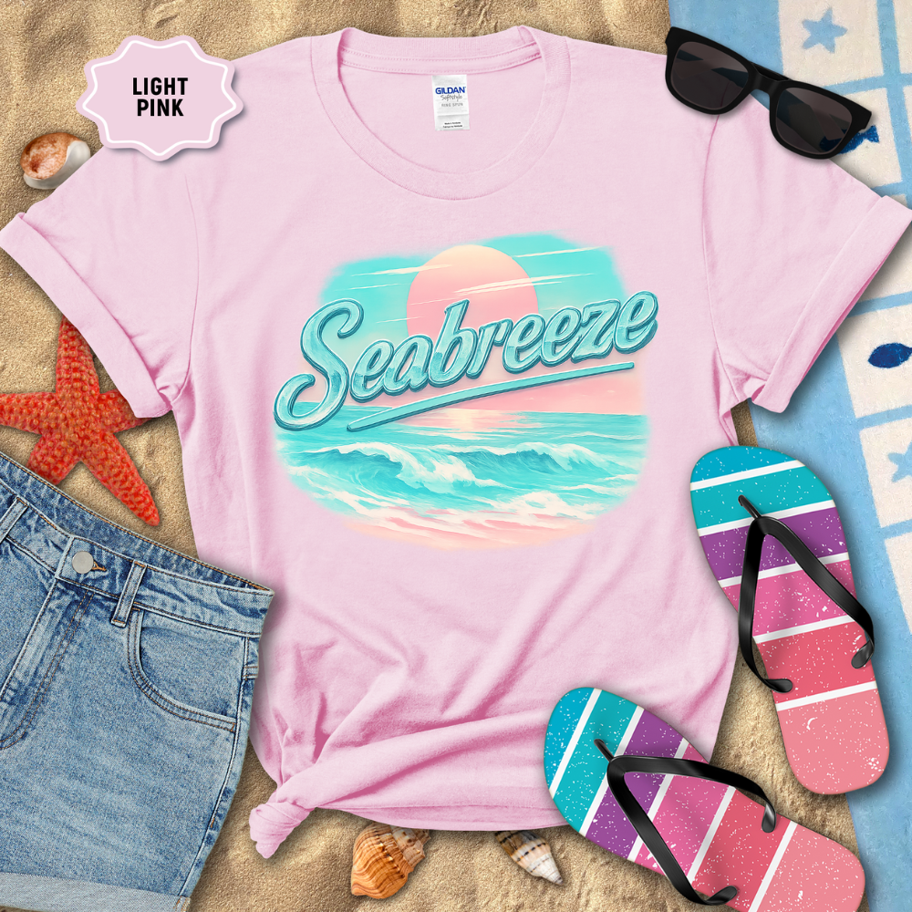 Light pink t-shirt with 'Seabreeze' design on a sandy surface with beach accessories.