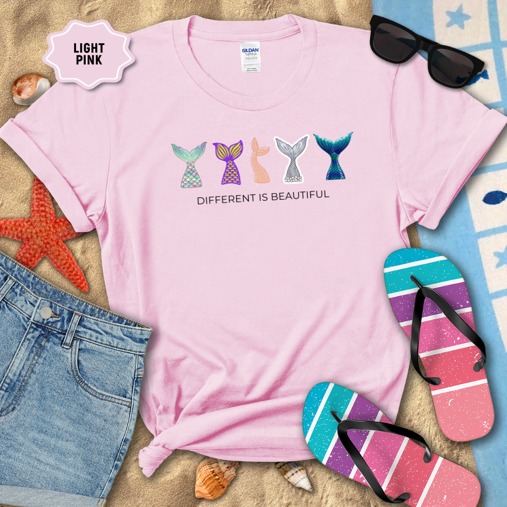 Light pink t-shirt with mermaid tail designs and 'Different is Beautiful' text, placed on sand with beach accessories.