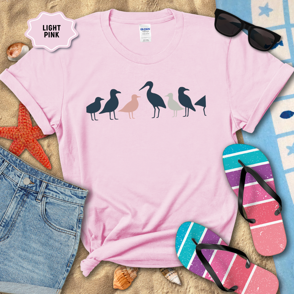 Light pink t-shirt with bird design on a sandy surface with beach accessories.