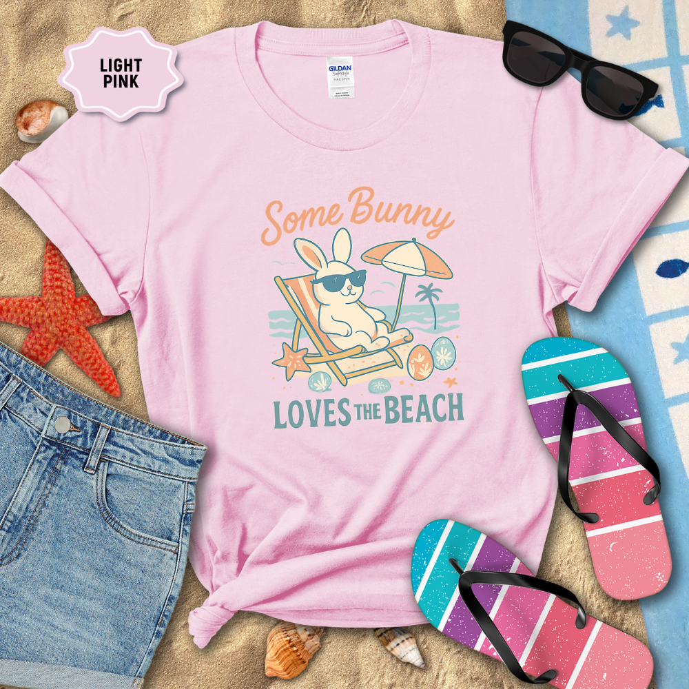 Light pink t-shirt with bunny design on a sandy background with flip-flops and sunglasses.