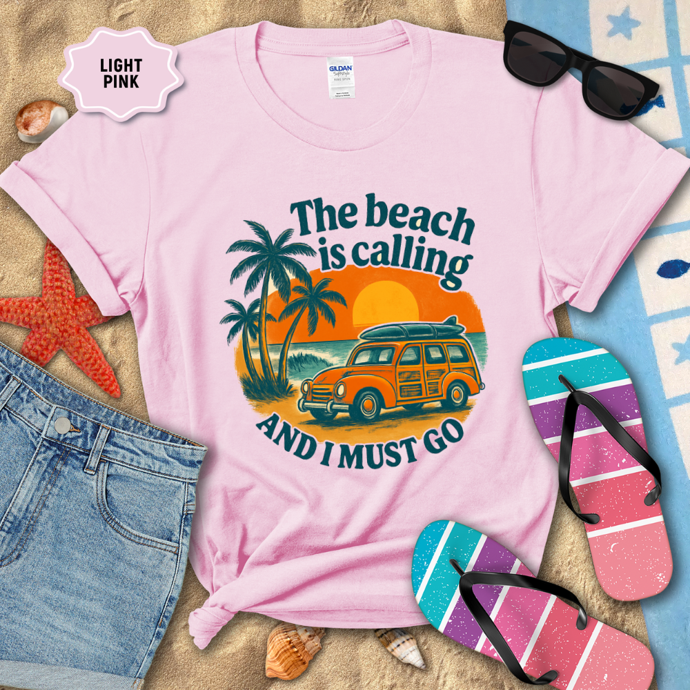 Light pink t-shirt with beach-themed design on sand with sunglasses, flip-flops, and denim shorts.