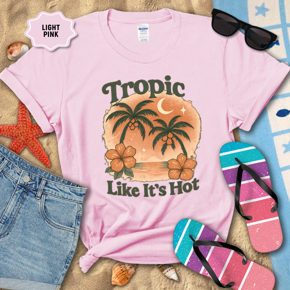 Light pink t-shirt with 'Tropic Like It's Hot' design, sunglasses, flip-flops, and beach items on sand.