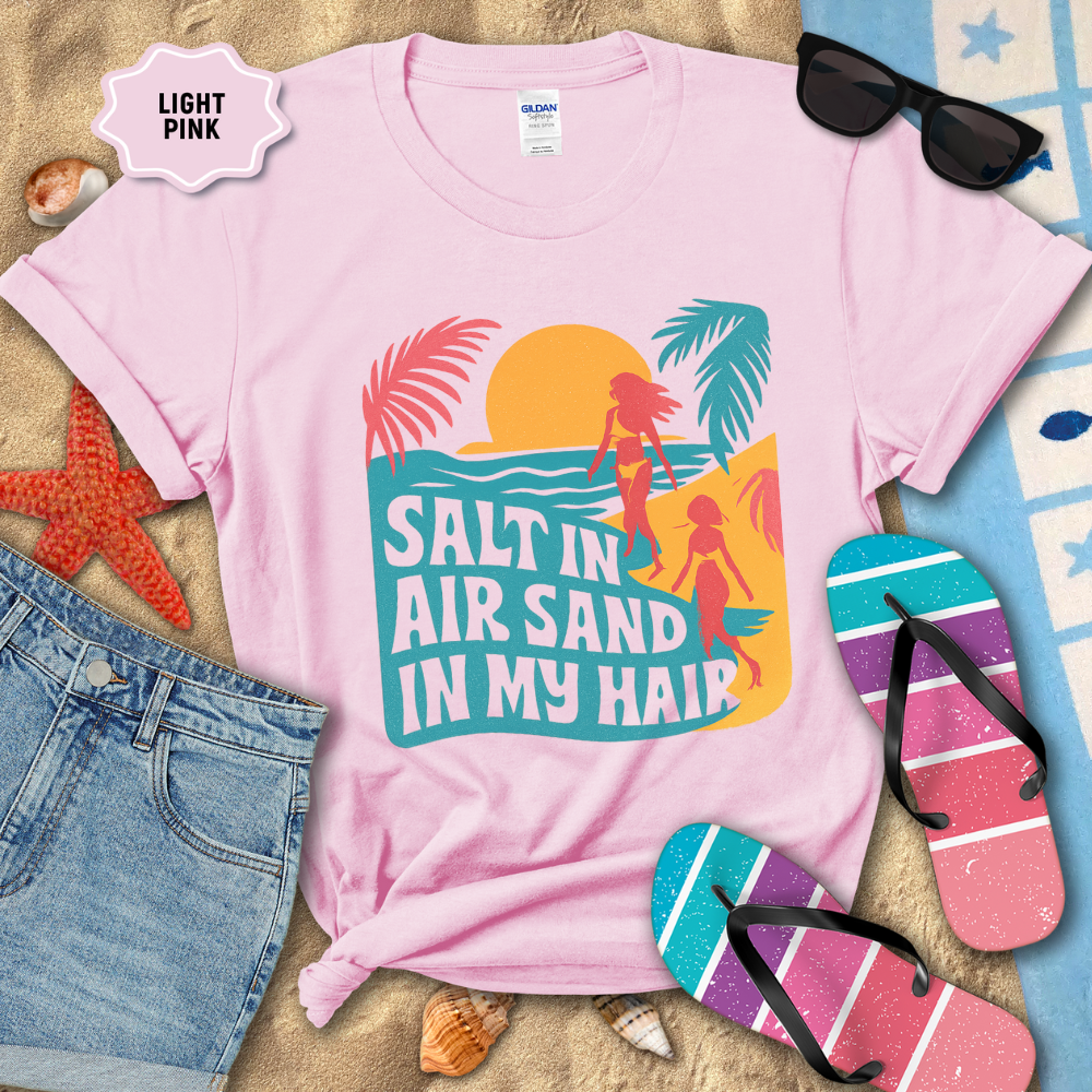 Light pink t-shirt with beach-themed design and text, surrounded by beach accessories on sand.