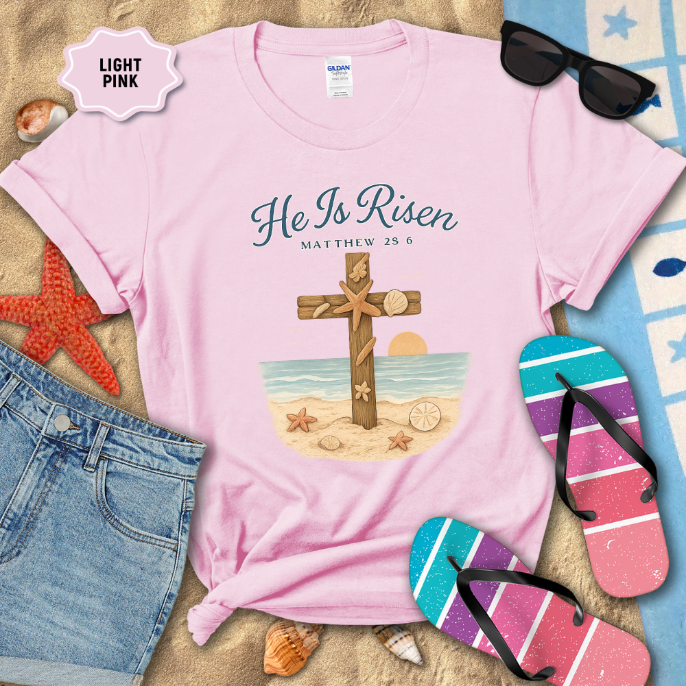 Light pink t-shirt with 'He Is Risen' design on a sandy background with flip-flops and sunglasses.