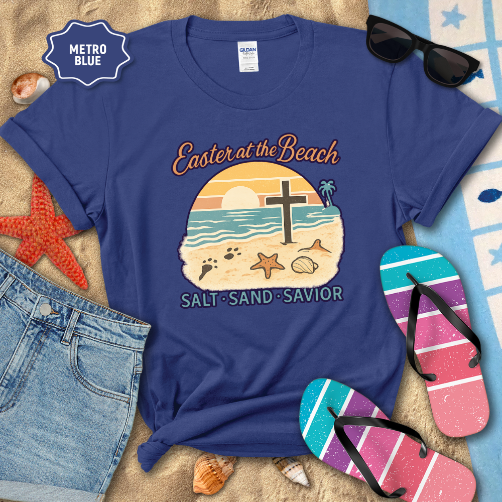 Blue t-shirt with beach-themed design on a sandy background with flip-flops and sunglasses.