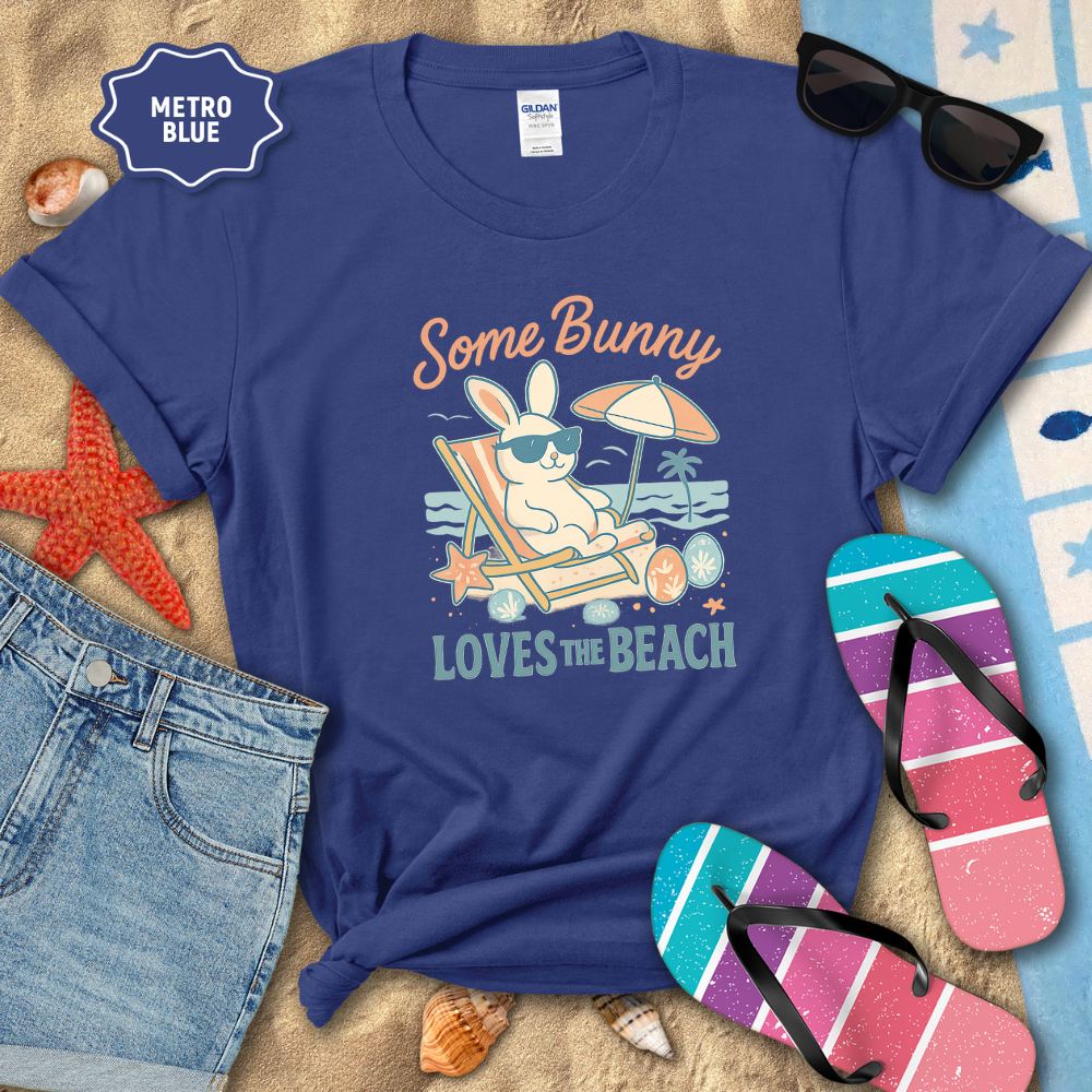 Blue t-shirt with beach-themed design featuring a bunny, sunglasses, and flip-flops on sand with beach accessories.